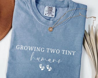 May include: Blue jean colored t-shirt with the text "GROWING TWO TINY humans" in white script. The shirt has a crew neck and short sleeves. A gold necklace with a pendant rests on the shirt.