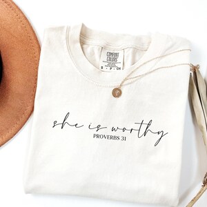 May include: Cream-colored t-shirt with the words "she is worthy" and "Proverbs 31" printed in black script. A gold necklace with a circular pendant rests on the shirt. A brown hat and dried plant are in the background.