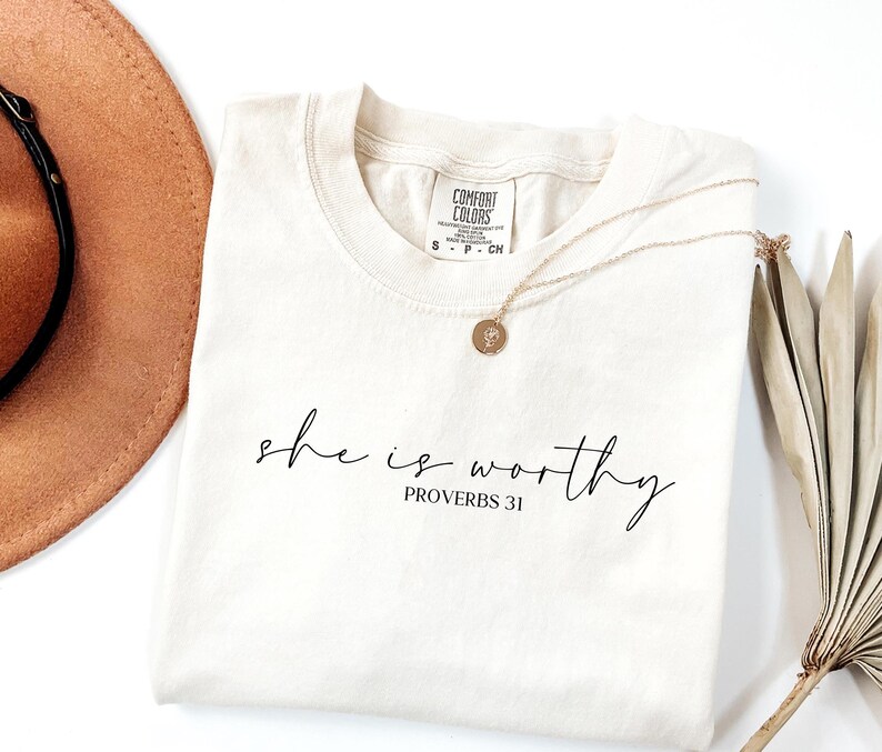 May include: Cream-colored t-shirt with the words "she is worthy" and "Proverbs 31" printed in black script. A gold necklace with a circular pendant rests on the shirt. A brown hat and dried plant are in the background.