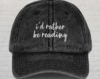 May include: A black distressed denim baseball cap with white embroidered text that reads "i'd rather be reading".