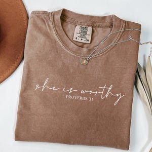 May include: A brown Comfort Colors t-shirt with the words "she is worthy" and "Proverbs 31" printed in white script. A silver necklace with a small pendant rests on the shirt. A brown hat and dried plant are in the background.