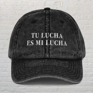 Può includere: Un cappellino da baseball nero in stile vintage con la scritta ricamata "TU LUCHA ES MI LUCHA" in bianco. Il cappellino ha una visiera curva e un bottone sulla parte superiore. Il tessuto ha un aspetto usurato e sbiadito.