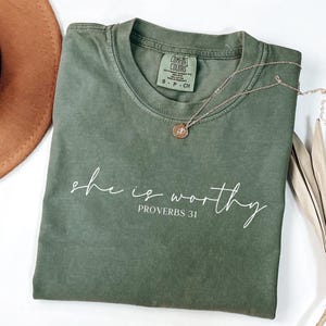 May include: An olive green t-shirt with the words "she is worthy" and "Proverbs 31" printed in white script. A gold necklace with a pendant and a brown hat are also in the image.