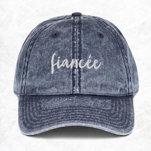Puede incluir: Una gorra de béisbol de mezclilla azul con la palabra "fiancée" bordada en blanco en la parte delantera.