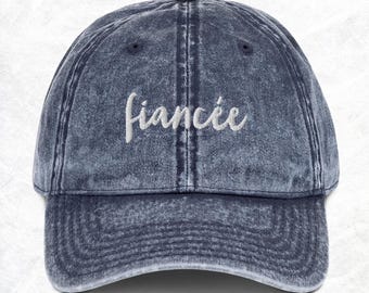 May include: A blue denim baseball cap with the word "fiancée" embroidered in white on the front.