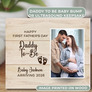 May include: Wooden photo frame with a picture of a couple, and text that reads "Happy First Father's Day Daddy To-Be" with baby footprints and the name "Baby Jackson Arriving 2026".