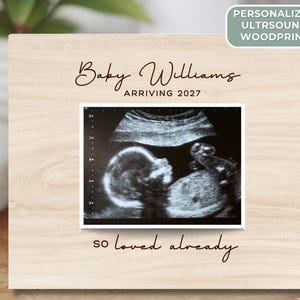 May include: A wooden frame with a sonogram image of a baby. The frame has the words "Baby Williams ARRIVING 2027" and "so loved already" in script. A green label reads "PERSONALIZED ULTRSOUND WOODPRINT".