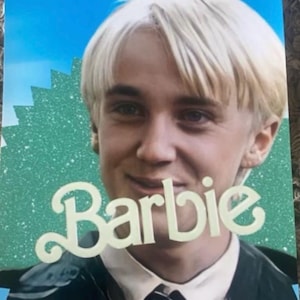 May include: Poster featuring a man with blonde hair, smiling, with the word "Barbie" in a cursive font overlaid on the image. The text "This Barbie is A natural blonde" is at the top. The background is blue and green.