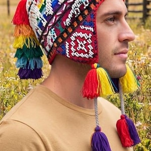 May include: A knitted earflap hat featuring a multi-coloured geometric pattern with white, red, blue, and green accents. The hat includes a large multi-coloured tassel on the back and two braided cords with smaller multi-coloured tassels on the sides.