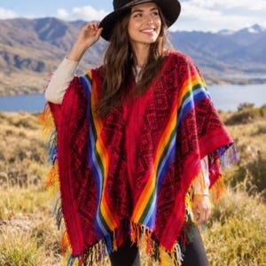 Puede incluir: Un poncho rojo con un patrón geométrico negro y rayas verticales de arcoíris en la parte delantera. La prenda tiene un dobladillo con flecos y borlas multicolores. Se lleva un sombrero negro de ala ancha con una banda decorativa.