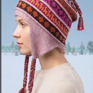 May include: A knit earflap hat featuring a multi-colored geometric pattern in shades of pink, orange, brown, and purple. The hat includes a solid light pink ribbed brim, earflaps, and three braided tassels with multi-colored yarn ends.