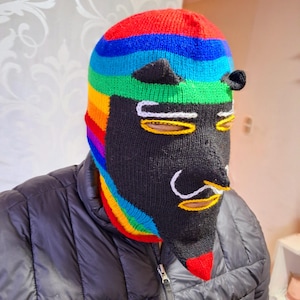 May include: A black knitted balaclava with a rainbow stripe pattern and a cartoon face design. The face has white eyebrows, yellow eyes, and a white mustache.