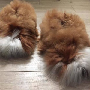 May include: A pair of fluffy slippers featuring a thick, long-pile texture in a warm brown color. Each slipper has a distinct patch of white fur at the toe area, creating a contrasting visual element against the brown base of the footwear.