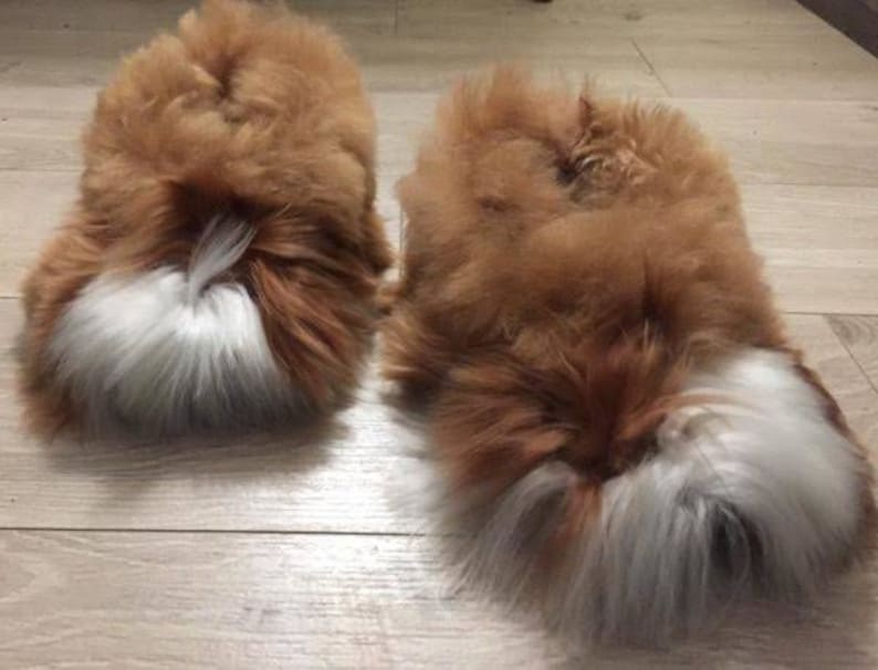 May include: A pair of fluffy slippers featuring a thick, long-pile texture in a warm brown color. Each slipper has a distinct patch of white fur at the toe area, creating a contrasting visual element against the brown base of the footwear.