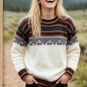 May include: A long-sleeved knit jumper featuring a cream-coloured lower section and a patterned upper yoke. The yoke displays horizontal bands of brown, teal, and black, including a repeating geometric mountain-like motif across the chest.