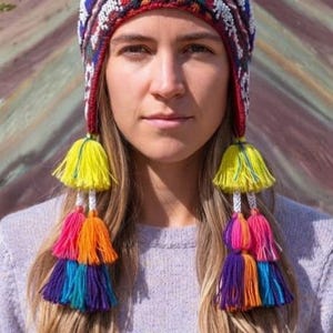 Könnte beinhalten: Eine gestrickte Chullo-Mütze mit einem lebendigen, mehrfarbigen geometrischen Muster und weißen Lamamotiven. Die Mütze ist mit einem Regenbogen-Bommel versehen und hat zwei lange geflochtene Ohrenklappen, die mit gelben, pinken, orangen, blauen und violetten Quasten enden.