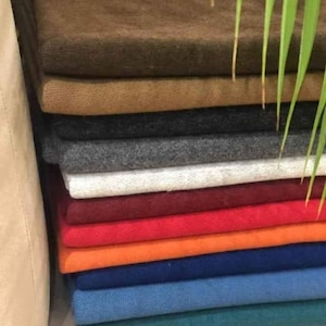 Puede incluir: Una pila de piezas de tela dobladas en varios colores sólidos, incluyendo marrón oscuro, bronceado, gris carbón, gris claro, blanco, granate, rojo, naranja, azul oscuro, azul medio y verde azulado. Cada tela tiene una superficie suave y texturizada.