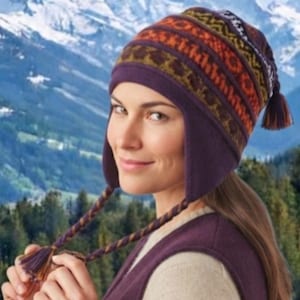 May include: A purple knitted hat with ear flaps and braided tassels. The hat features a multi-coloured patterned band in shades of orange, brown, and yellow. The design includes a solid purple base with matching ear flaps and long braided ties.