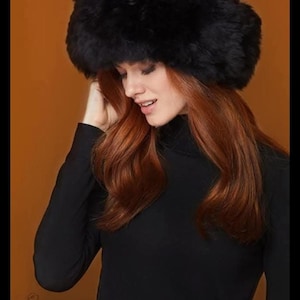 May include: A black faux fur hat with a thick, plush texture and a rounded crown. The hat features a wide, folded brim that encircles the head, providing full coverage. The material appears soft and dense, with a deep black color throughout.