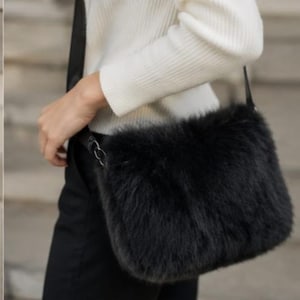 May include: A black faux fur shoulder bag with a rectangular shape and a long, thin black strap. The bag features a dense, fluffy texture and is attached to the strap with small black metal hardware rings on the sides of the main compartment.