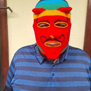 Puede incluir: Un pasamontañas tejido con un diseño de diablo. La parte superior del pasamontañas está a rayas con colores del arcoíris, y la cara es roja con ojos delineados en amarillo, un bigote y cuernos pequeños. La persona lleva un polo a rayas azules.