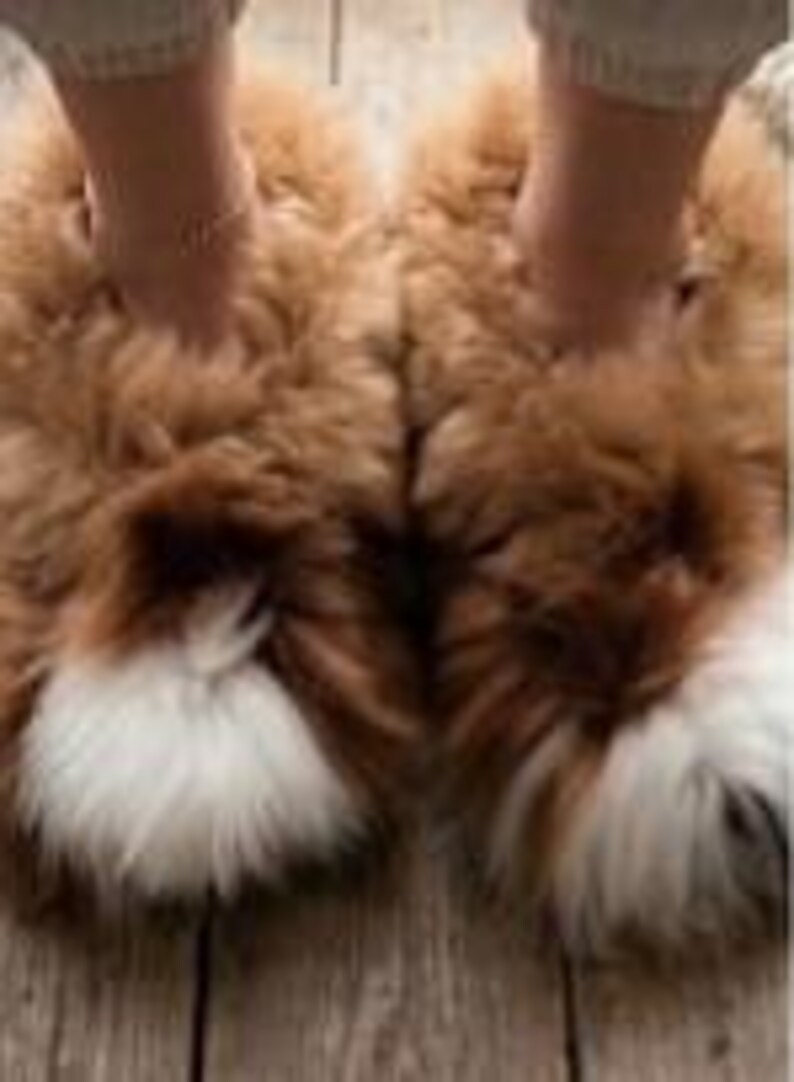 May include: A pair of fluffy, thick-pile slippers in a warm tan brown color, featuring a distinct patch of white fur at the rounded toe area. The slippers have a high-loft, textured appearance and are designed as slip-on footwear for indoor use.