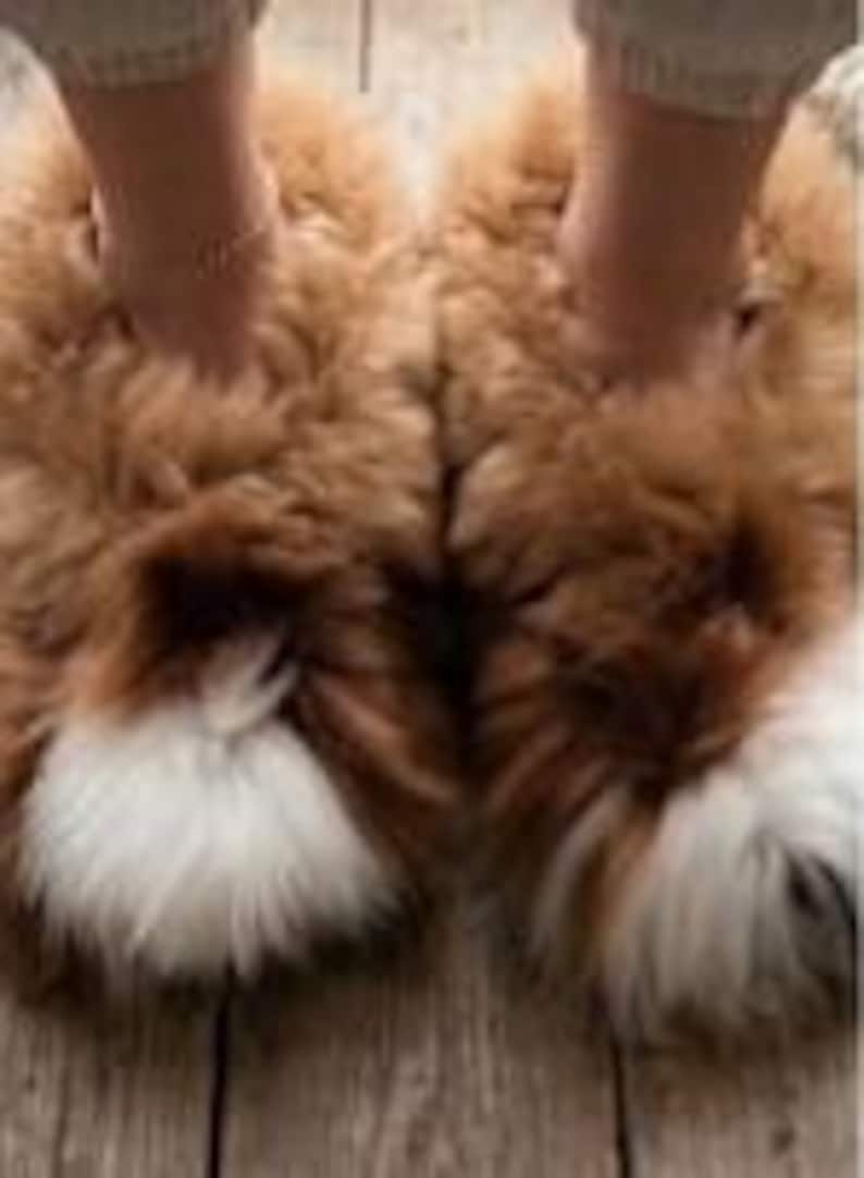 May include: A pair of fluffy, thick-pile slippers in a warm tan brown color, featuring a distinct patch of white fur at the rounded toe area. The slippers have a high-loft, textured appearance and are designed as slip-on footwear for indoor use.