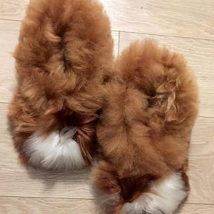 May include: A pair of fluffy slippers made from long-pile brown fur. Each slipper features a distinct patch of white fur at the toe area, creating a contrasting color detail against the primary brown texture of the footwear.