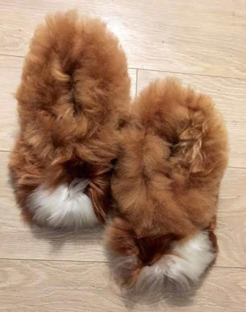 May include: A pair of fluffy slippers made from long-pile brown fur. Each slipper features a distinct patch of white fur at the toe area, creating a contrasting color detail against the primary brown texture of the footwear.