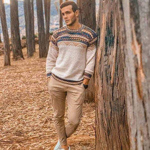 May include: A man wearing a white sweater with a colourful geometric pattern, beige trousers, and white trainers. He is standing in a forest with trees behind him.