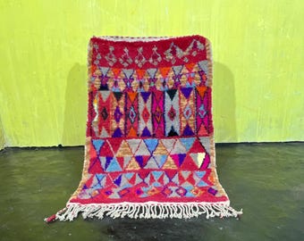 May include: A colorful Moroccan rug with geometric patterns in shades of red, pink, blue, orange, and yellow. The rug has a fringe border and is made of wool.
