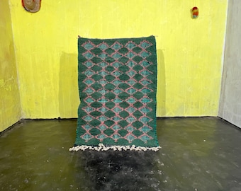 May include: A green and pink patterned rug with fringe on the bottom edge. The rug is hanging on a yellow wall.