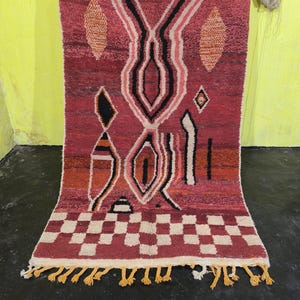 May include: A rectangular, hand-woven rug with a geometric design. The rug features a predominantly red background with black, white, and orange geometric shapes. The bottom edge has a checkered pattern and yellow tassels.