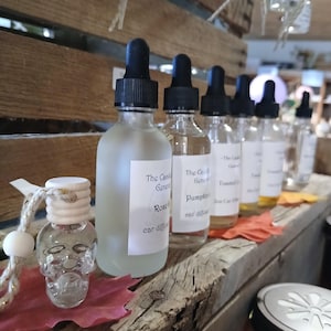 May include: A row of six small glass bottles with black droppers, filled with clear liquid, labeled with the text "The Candle General" and different scents, including "Rose & Cedar" and "Pumpkin Spice". The bottles are arranged on a wooden shelf with a small skull-shaped bottle hanging from a string in the foreground.