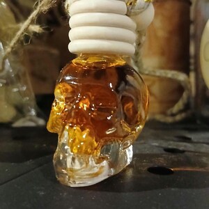 May include: A clear glass skull-shaped bottle filled with an amber-colored liquid. The bottle features a ribbed wooden cap and a hanging rope. A white paper tag is inserted into the top of the cap with the text Hocus Pocus printed on it.
