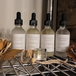 May include: Four clear glass bottles with black dropper tops and labels that read "The Candle Shop Geneva NY" and different scents: Lilac Gardens, Greenhouse, Magnolia & Peony, and Palo Santo. Each bottle has a label that says "car diffuser refill".