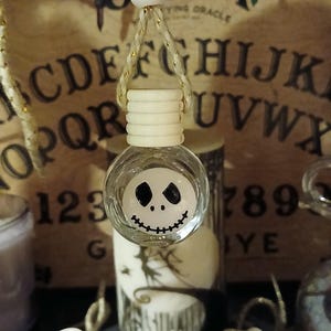 May include: A clear glass air freshener with a white lid and a string for hanging. The air freshener has a painted face of Jack Skellington from The Nightmare Before Christmas. Other similar air fresheners are visible in the background.