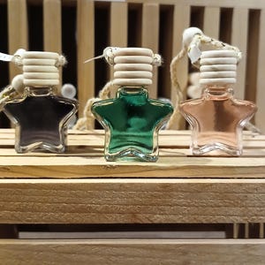 May include: Three star-shaped car air fresheners in black, green, and peach. Each has a wooden top and a string for hanging. They are displayed on a wooden shelf, with other products visible in the background.
