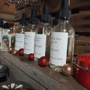 May include: Four clear glass bottles with droppers filled with scented oil. The bottles have white labels with black text that reads "Candle Shop - Geneva NY" and the scent name. The bottles are arranged on a wooden shelf with red and gold decorative balls.