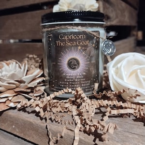 May include: A black and white candle jar with a label that reads "Capricorn The Sea Goat". The label also includes the astrological symbol for Capricorn, a goat's head, and the text "Element: Earth, Color: Grey, Stone: Garnet, Ruling Planet: Saturn, Spirit Animal: Goose, December 22nd - January 19th".