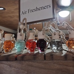 May include: Seven glass skull-shaped car air fresheners hanging from strings. The scents are Cinnamon, Violet Star, and five other scents. The air fresheners are displayed on a wooden surface with a sign that reads "Air Fresheners".