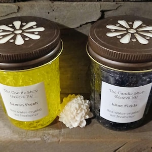 May include: Two glass jars with brown lids and white flower designs. The jars contain yellow and black water crystals. The jars are labeled "The Candle Shop Geneva NY" and have the scents "lemon Fresh" and "lilac Fields".