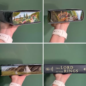 Pu&ograve; includere: Una copia con copertina rigida di The Lord of the Rings di J.R.R. Tolkien con copertine nere e scritte dorate. I bordi delle pagine presentano scene dipinte a mano, tra cui un paesaggio verde con una porta rotonda, un edificio e delle cascate.