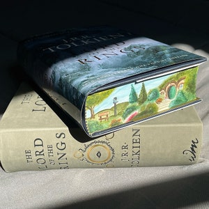 Pu&ograve; includere: Una copia con copertina rigida dell'edizione del 50&deg; anniversario de Il Signore degli Anelli di J.R.R. Tolkien, con un'illustrazione dipinta a mano sul taglio delle pagine che raffigura una tana di hobbit verde, alberi e una panchina.
