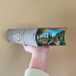 Pu&ograve; includere: Un libro con copertina rigida grigia intitolato The Lord of the Rings di J.R.R. Tolkien con un paesaggio dipinto a mano sul bordo delle pagine. L'opera raffigura una collina verde con una porta rotonda, alberi, una panchina e una casetta.