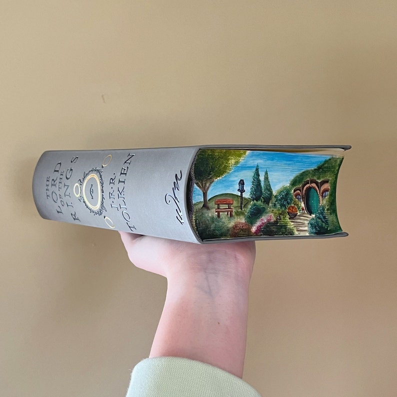 Pu&ograve; includere: Un libro con copertina rigida grigia intitolato The Lord of the Rings di J.R.R. Tolkien con un paesaggio dipinto a mano sul bordo delle pagine. L'opera raffigura una collina verde con una porta rotonda, alberi, una panchina e una casetta.