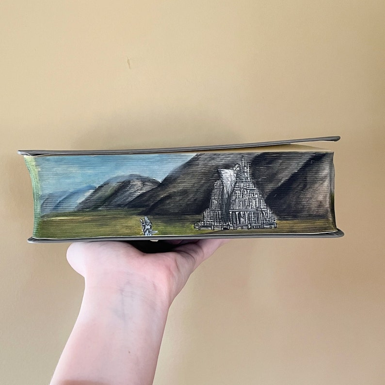 Pu&ograve; includere: Un libro con pittura sul taglio anteriore delle pagine, raffigurante un'illustrazione paesaggistica dettagliata di una struttura alta a pi&ugrave; livelli, simile a una torre o un tempio, contro montagne scure e ondulate sotto un cielo blu con una piccola figura.