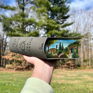 Pu&ograve; includere: Un libro con copertina rigida grigia intitolato The Lord of the Rings di J.R.R. Tolkien con una scena dipinta a mano sui bordi delle pagine che raffigura una porta circolare verde, una panchina in legno, alberi e cespugli in un paesaggio.