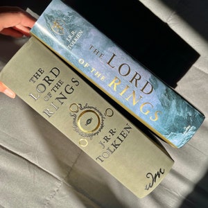 Pu&ograve; includere: Due libri con copertina rigida intitolati The Lord of the Rings di J.R.R. Tolkien. Un libro presenta una copertina verde oliva opaca con un simbolo dell'occhio dorato, l'altro una sovraccoperta paesaggistica blu e grigia con scritte dorate.