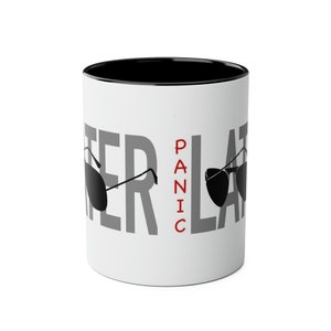 Puede incluir: Taza de café de cerámica blanca con borde negro. La taza tiene un gráfico gris que dice "PANIC LATER" con gafas de sol a cada lado del texto. La palabra "PANIC" está en rojo.